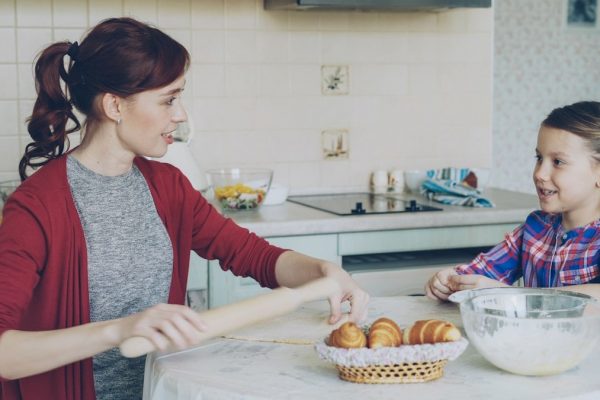 Young mother rolling dough and talking to her little cute daughter while cooking in the kitchen on weekend. Family, food, home and people concept